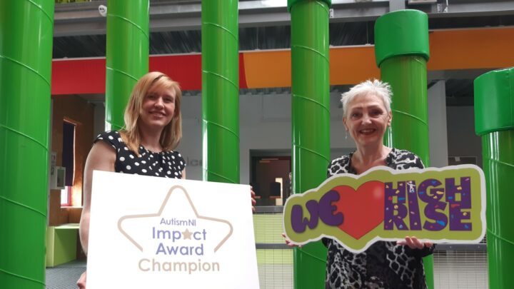 Christine Kearney, Autism NI presenting Marie Marin, Chief Executive Employers For Childcare with the Autism NI Impact Award on behalf of High Rise. To the left of the image, Christine is holding a large white sign with the Impact NI Impact Award Champion logo, Marie is standing to the right holding a sign reading 'We heart High Rise'.