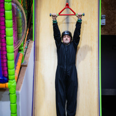 Male team member hanging on while being elevated on Ireland's tallest vertical drop slide during a team building session at High Rise Lisburn. He is wearing a safety helmet and protective jumpsuit.
